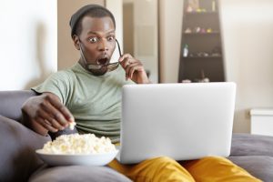 Surprised African male sitting on couch at home, eating popcorn and watching exciting TV show online on laptop computer or shocked with cliffhanger ending of detective series, keeping his mouth open